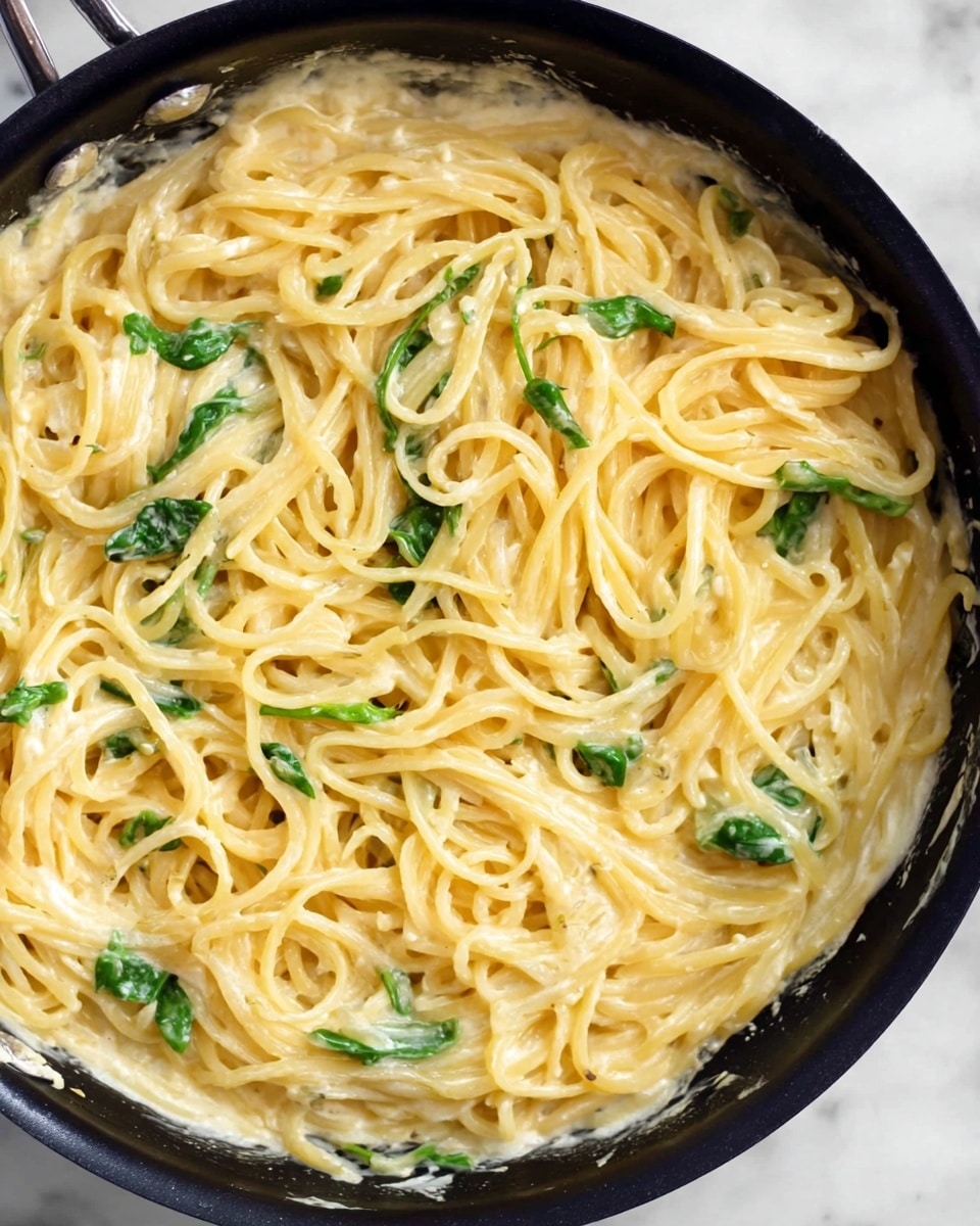 A black pan filled with creamy spaghetti pasta mixed in a smooth, light yellow sauce with small green spinach leaves scattered throughout. The pasta strands are thick and coated evenly in the sauce, showing a rich texture, with some bits of sauce splashed on the sides of the pan. The pan sits on a white marbled surface. photo taken with an iphone --ar 4:5 --v 7