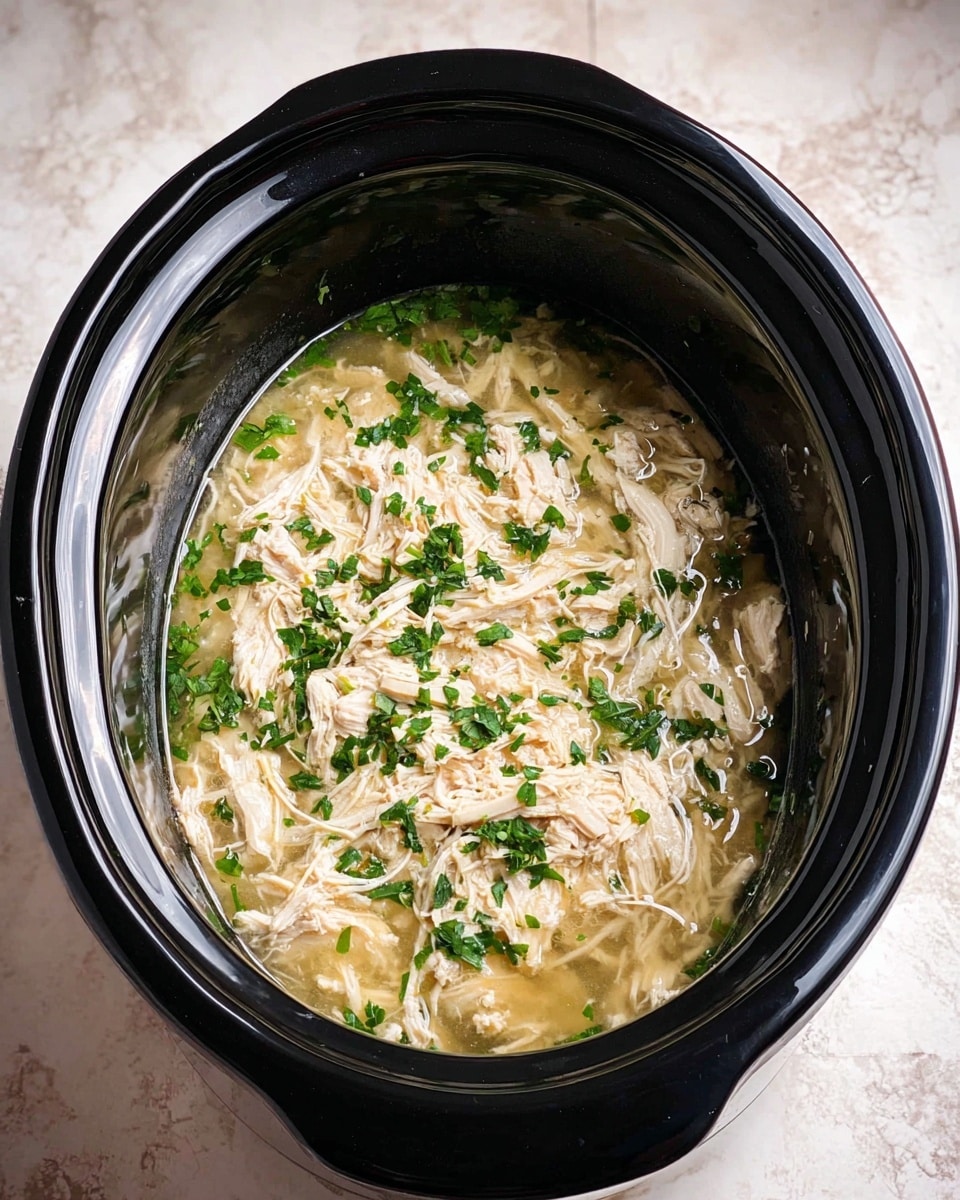 Inside a black slow cooker, there is one layer of shredded light beige chicken soaked in a clear broth and sprinkled with small, bright green parsley pieces. The chicken looks soft and moist, floating in the broth that fills the cooker almost halfway. The slow cooker is placed on a white marbled surface. Photo taken with an iphone --ar 4:5 --v 7