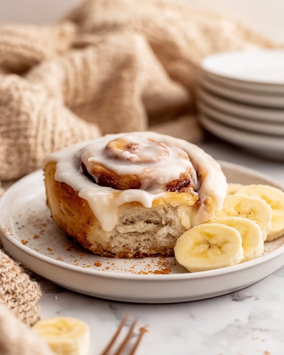 A close-up of a cinnamon roll with creamy white icing on top showing swirled layers of golden brown dough and cinnamon filling, placed slightly off-center on a white plate with two thick banana slices next to it, crumbs scattered around the roll on the plate, with a stack of clean white plates and a soft beige knitted cloth in the blurry background, all sitting on a white marbled surface photo taken with an iphone --ar 4:5 --v 7