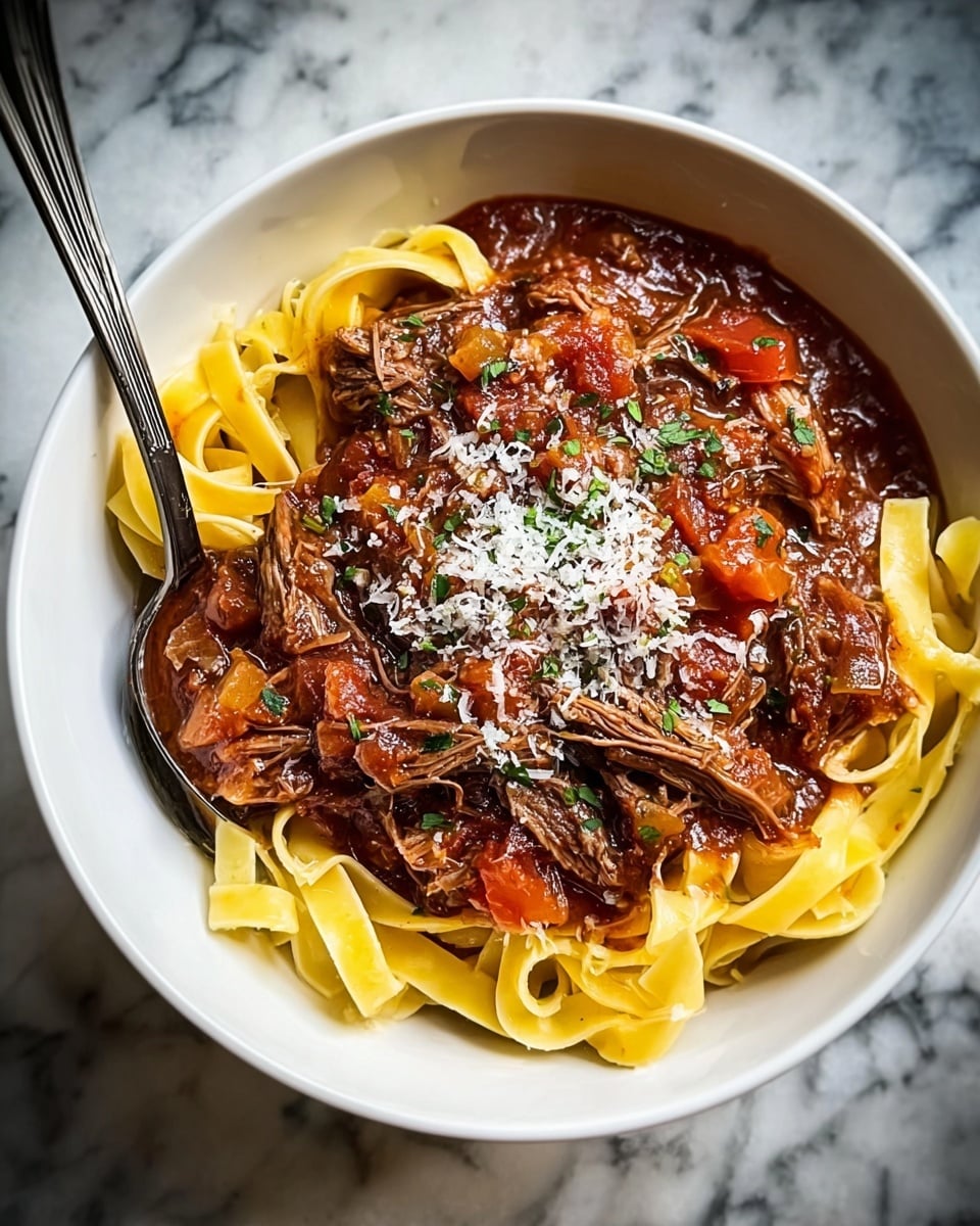 A white bowl filled with three main layers: at the bottom are wide, flat yellow pasta noodles arranged loosely, on top is a thick rich red-brown stew with visible shredded dark brown meat, chunks of red tomatoes, and pieces of translucent cooked onions, sprinkled over with small green herb bits and white grated cheese. The bowl rests on a white marbled surface, and a spoon is placed inside the bowl on the left side. photo taken with an iphone --ar 4:5 --v 7