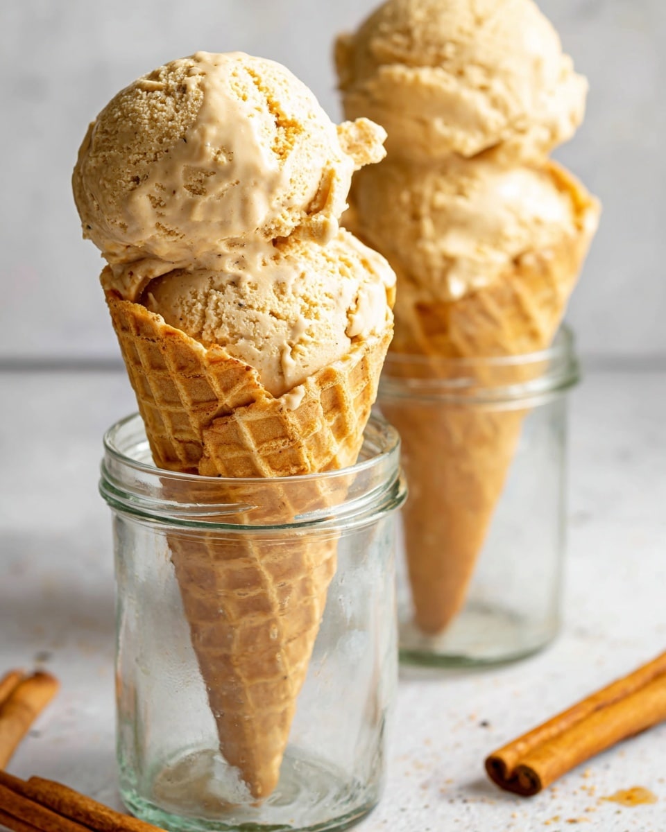 The image shows two scoops of light brown ice cream with a slightly rough texture, stacked on top of a waffle cone. The cone is placed inside a clear glass jar and has a light golden color with a classic waffle pattern. In the background, there is a white ceramic vase holding orange and yellow dried flowers and a white pumpkin. Two cinnamon sticks lie on a white marbled surface near the jar, and a small glass jar with a cork lid and brown powder is blurred in the background. Photo taken with an iphone --ar 4:5 --v 7