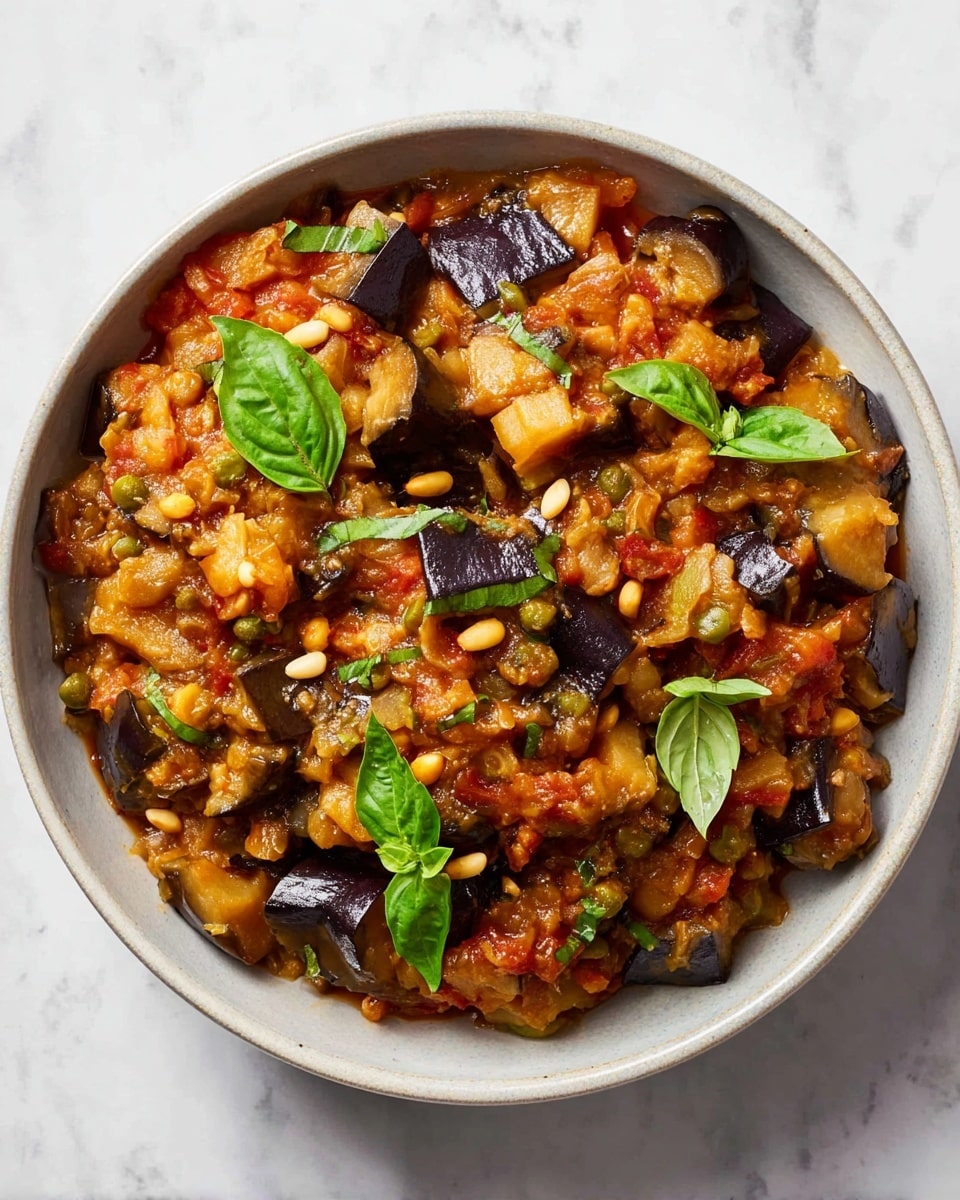 The image shows a white bowl filled with a chunky vegetable stew made of deep purple eggplant pieces, diced red tomatoes, small yellow pine nuts, and finely chopped yellow onions, all mixed in a rich reddish-orange sauce. Bright green fresh basil leaves are scattered on top, adding a pop of color. A silver spoon rests in the bowl, partially covered by the stew. The bowl sits on a white marbled surface. photo taken with an iphone --ar 4:5 --v 7