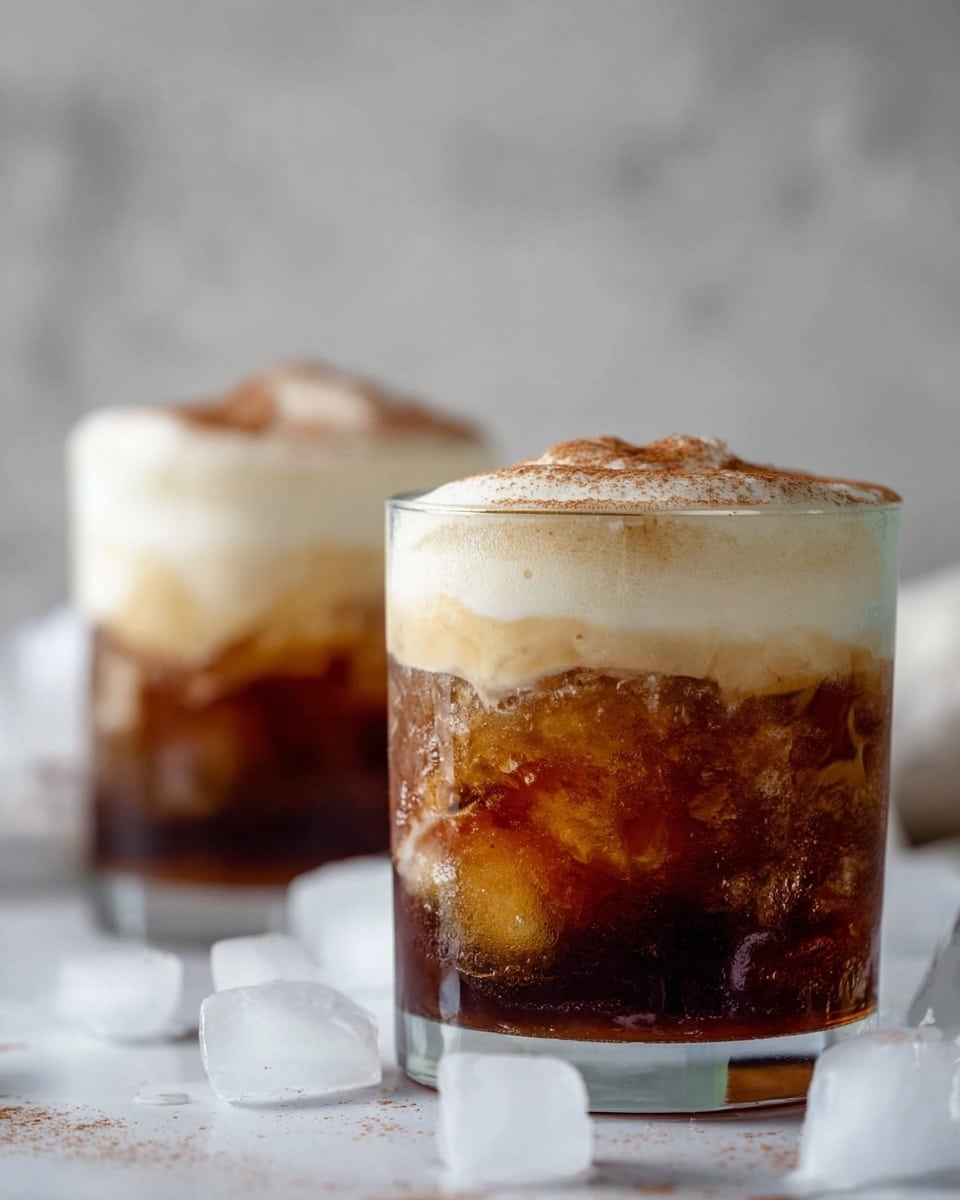 Two clear glasses sit on a white marbled surface with scattered ice cubes around them. Each glass shows three layers: the bottom is filled with crushed ice in dark brown liquid with amber highlights, the middle layer is creamy and light beige with a smooth texture, and the top layer is thick white foam sprinkled lightly with brown powder. The background is soft and blurred in neutral tones, putting focus on the drinks in the foreground. Photo taken with an iphone --ar 4:5 --v 7