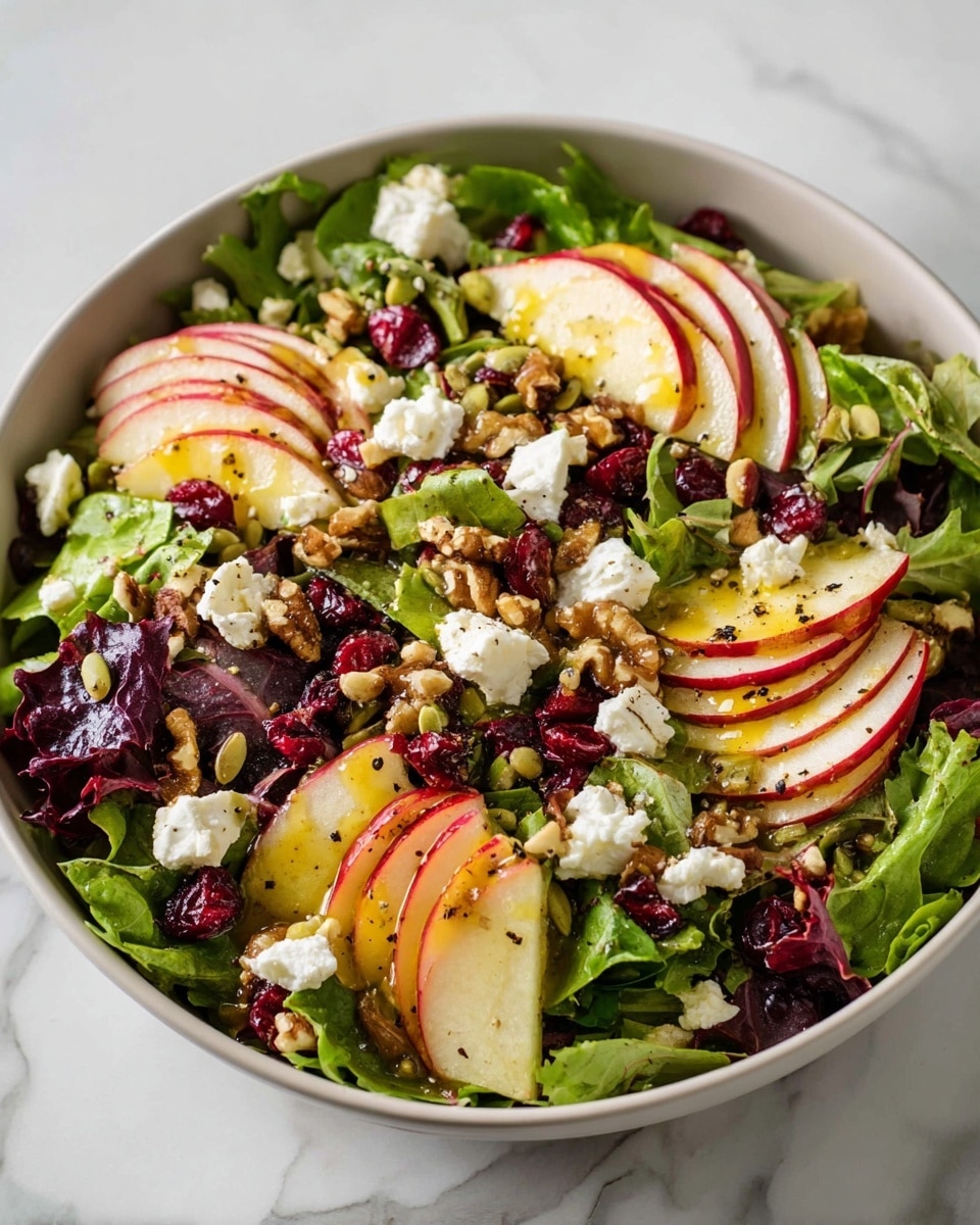 The dish shows a fresh mixed green salad with layers of leafy greens including light green and dark purple lettuce spread evenly. Thin, bright red apple slices with white flesh are arranged over the greens in curved rows. Small white crumbles of cheese, likely goat cheese, sit scattered on top. Dark red dried cranberries are sprinkled across the salad, along with small brown pumpkin seeds and broken walnut pieces in light brown. A light drizzle of dressing gives a slight shine to the ingredients. Everything rests on a white marbled surface photo taken with an iphone --ar 4:5 --v 7