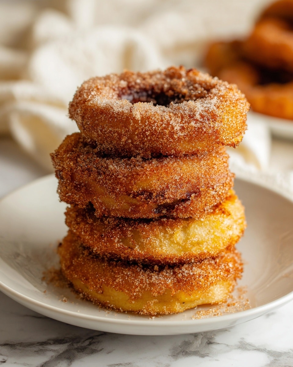 A stack of five round, golden apple rings covered in a crumbly cinnamon sugar coating sits in the center of a white plate, each ring showing a rough, textured surface with crispy edges. The top layer has a slightly irregular shape with visible sugar granules sparkling on it, while the inner layers reveal a softer, yellowish apple flesh beneath the coating. The plate rests on a white marbled surface, with a blurred white cloth and more apple rings in the background. photo taken with an iphone --ar 4:5 --v 7